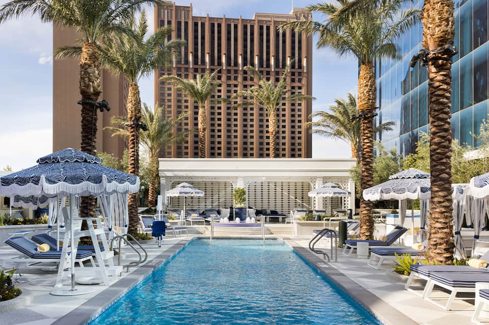Modern hotel pool with blue umbrellas and lounge chairs, surrounded by tall buildings and palm trees
