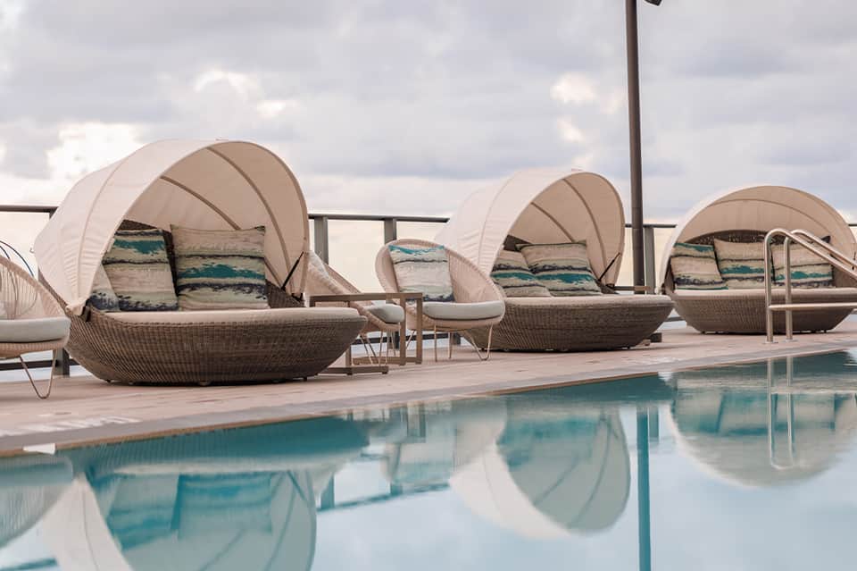 Three woven lounge chairs with canopies and striped cushions beside a calm turquoise pool