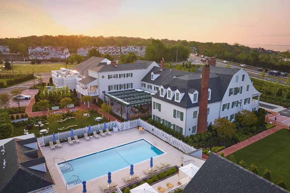 Aerial view of white New England-style resort with pool, lounge chairs, and surrounding residential buildings at dusk