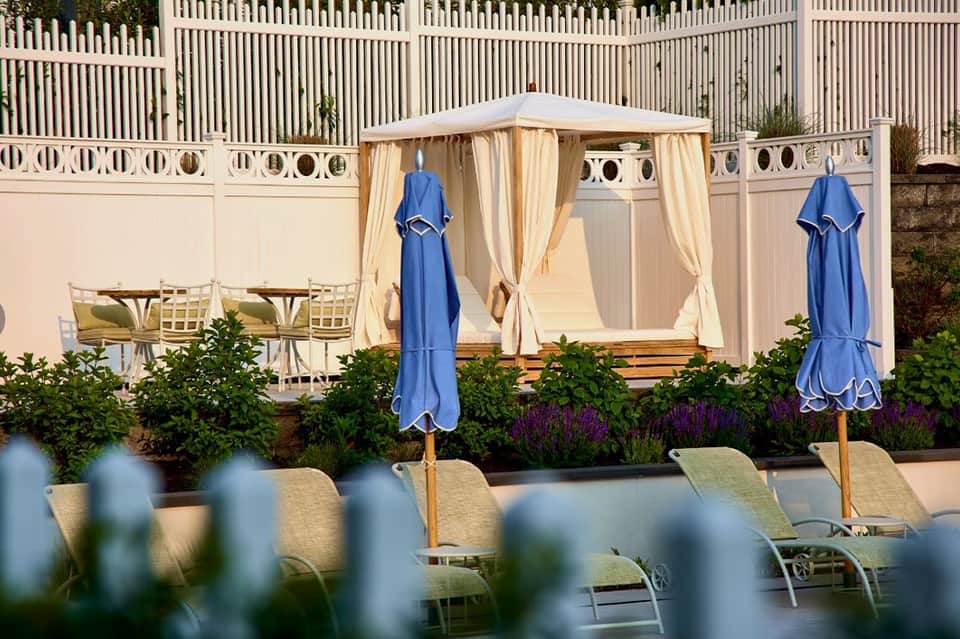 Elegant poolside cabana with cream curtains, blue umbrellas, lounge chairs, and manicured garden landscaping