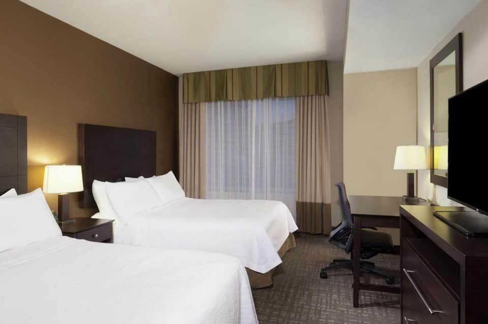 Modern hotel room with two queen beds, dark headboard, work desk, and floor-to-ceiling windows with curtains