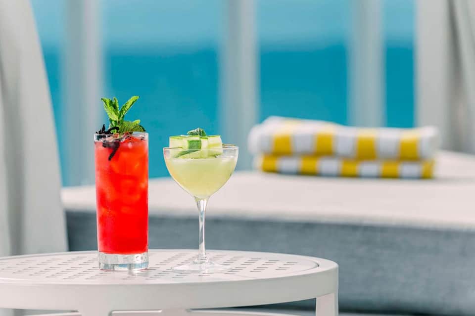 Two colorful cocktails on a white table with a turquoise ocean view and pool lounge in the background