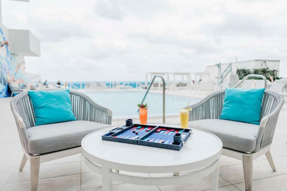 Two white modern chairs with turquoise pillows facing round table with backgammon board on beach terrace