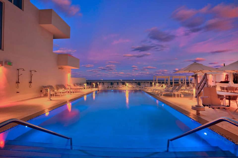 Illuminated resort pool at twilight with lounge chairs, umbrellas, and pink building facade
