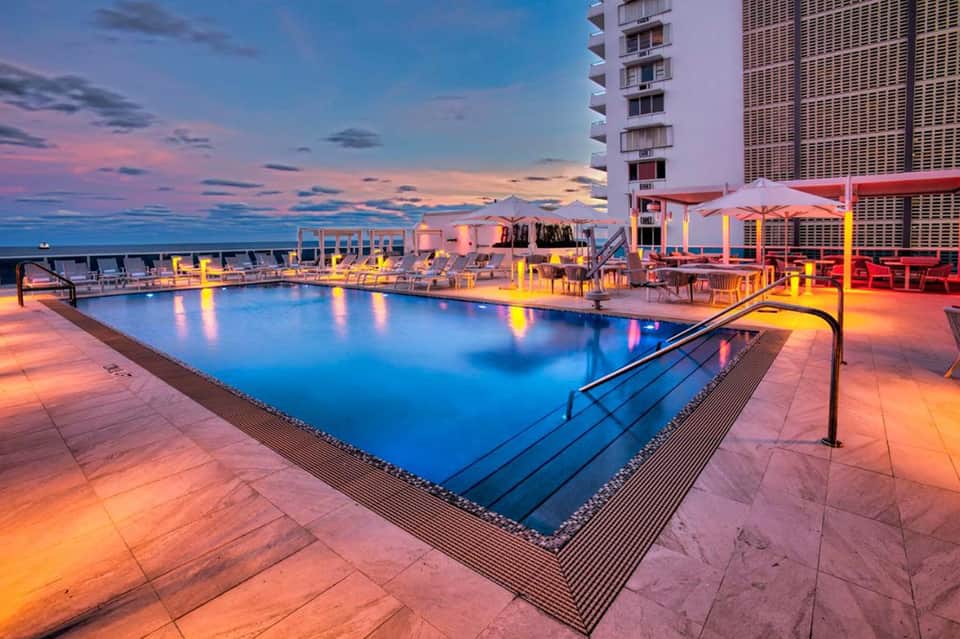 Resort pool at sunset with illuminated deck, umbrellas, and high-rise hotel buildings