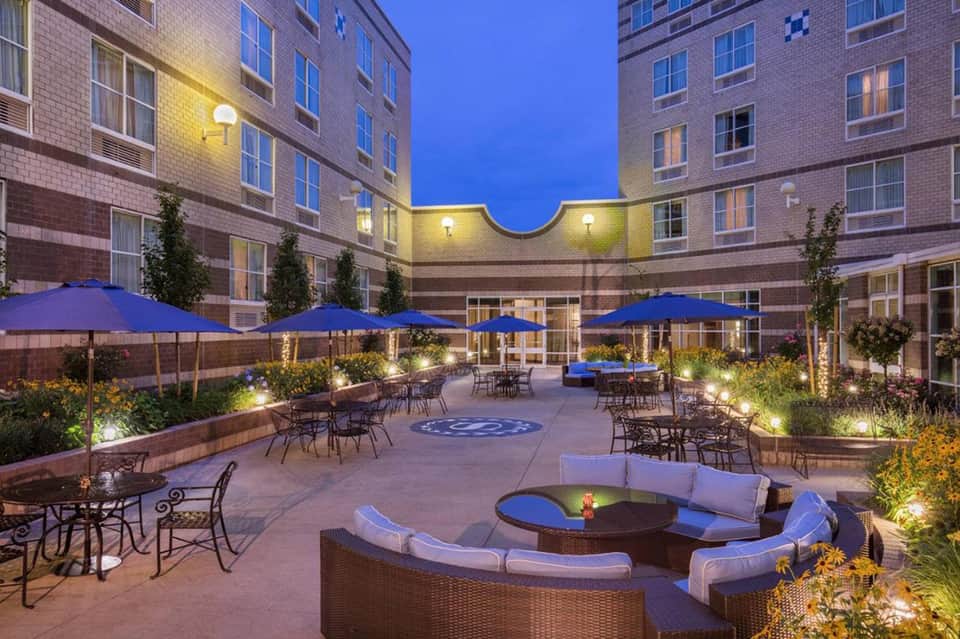 Illuminated courtyard with blue umbrellas, fire pit seating, and surrounding brick buildings at dusk
