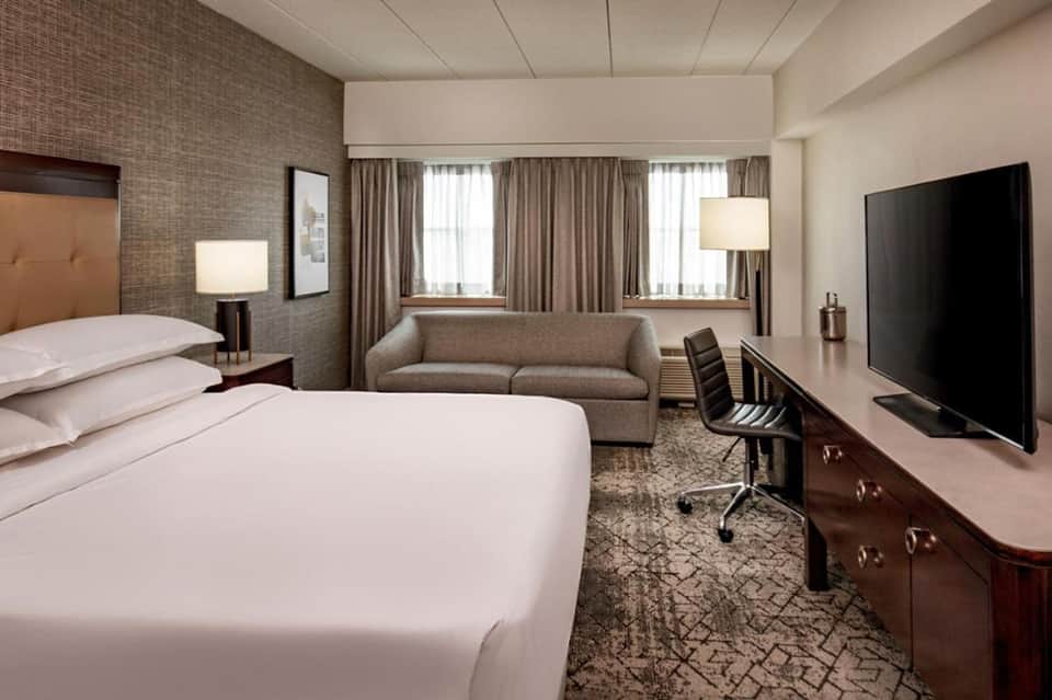 Modern hotel room with queen bed, work desk, sofa, and flat-screen TV in neutral tones