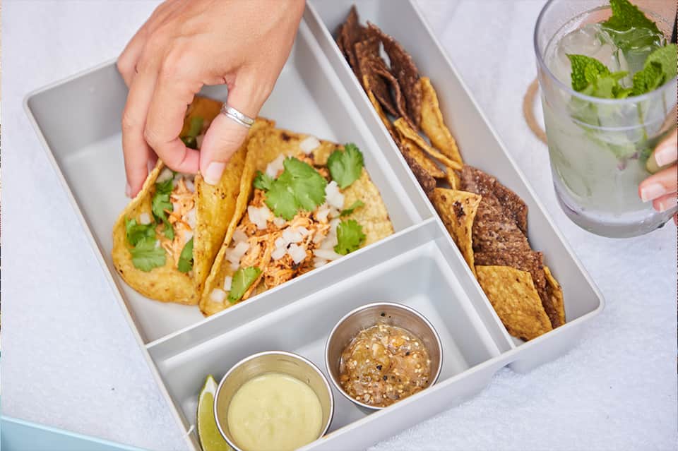 Plated tacos with shredded chicken, cilantro, and onions served with salsa and lime in a compartmentalized box