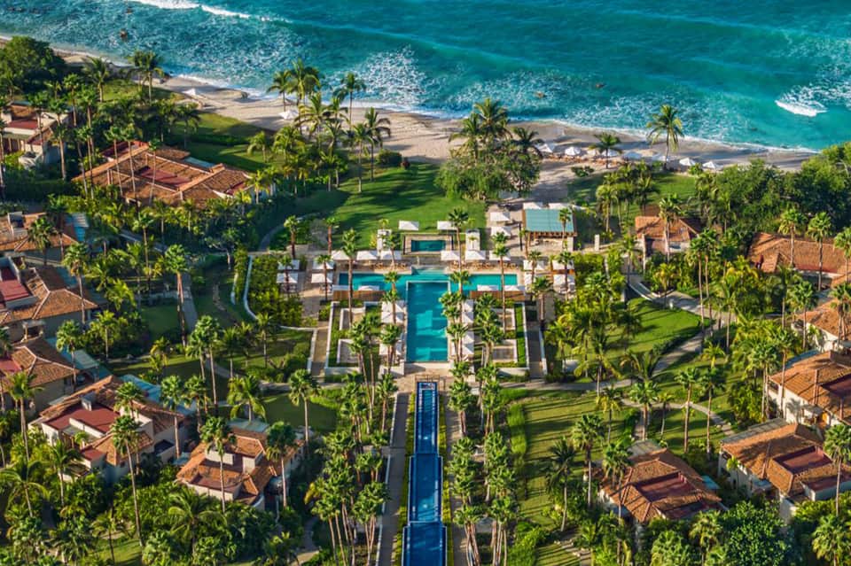Aerial view of beachfront luxury resort with central pool, palm trees, and ocean backdrop