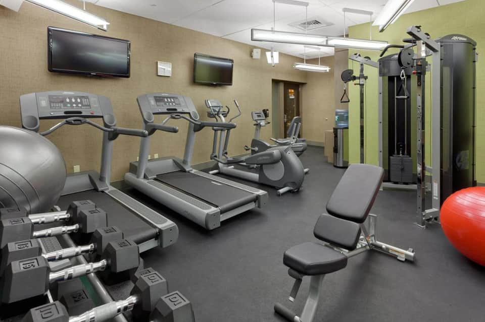 Modern fitness center with treadmills, exercise equipment, dumbbells, and TV screens on beige walls