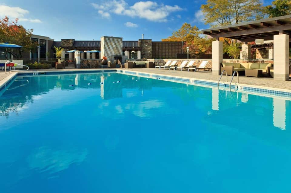 Resort swimming pool with blue water, lounge chairs, pergola, and stone building with autumn foliage