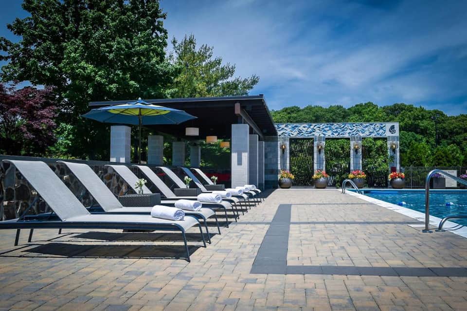 Resort pool deck with striped lounge chairs, blue umbrella, modern pavilion, and manicured gardens