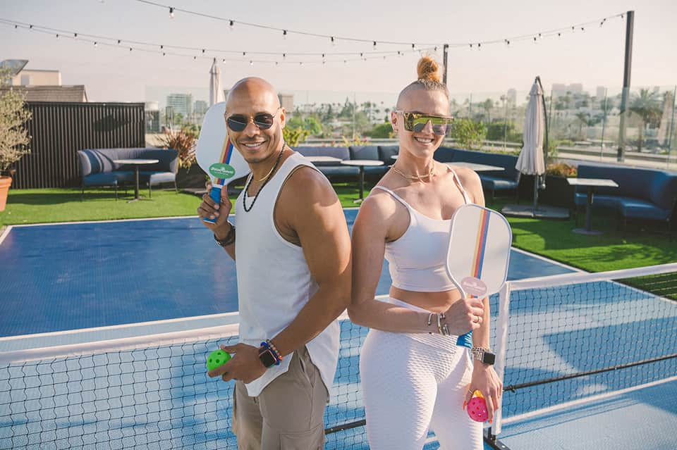 Couple holding pickleball paddles at rooftop pool with city skyline background