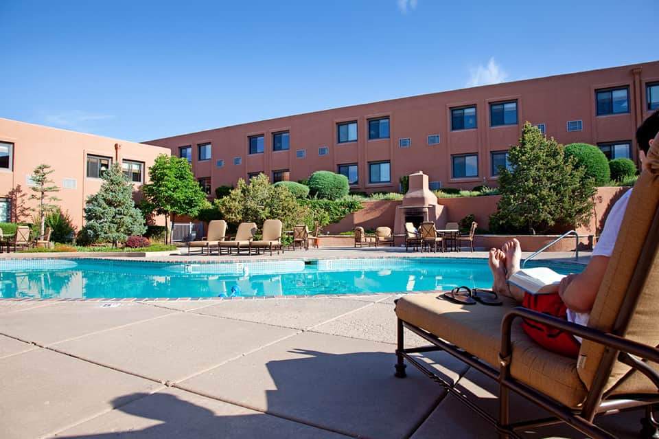 Resort guest relaxing on lounge chair by rectangular pool with red brick building and landscaping