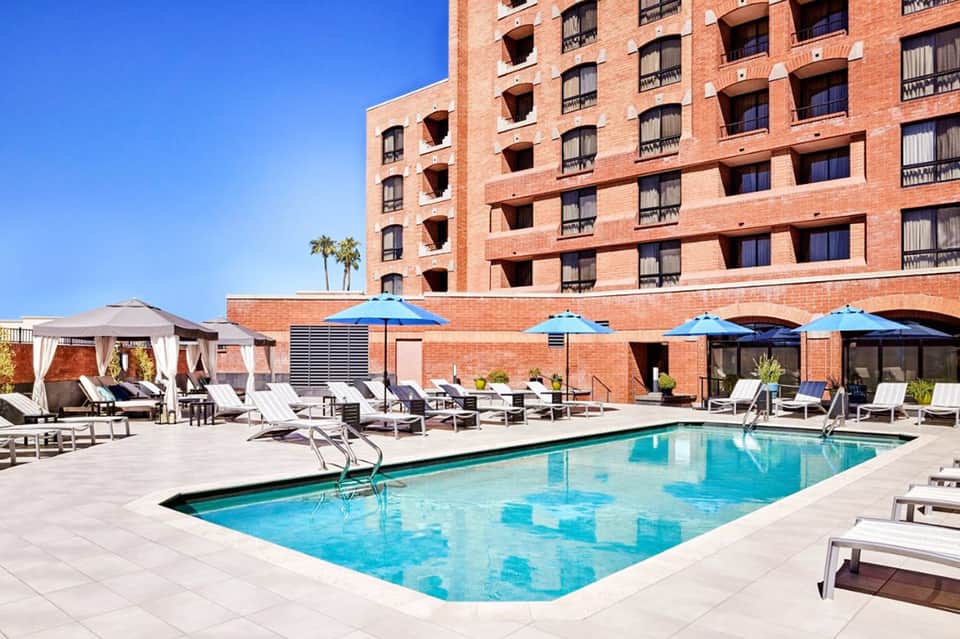 Brick hotel building with blue umbrellas and lounge chairs surrounding rectangular swimming pool under clear sky