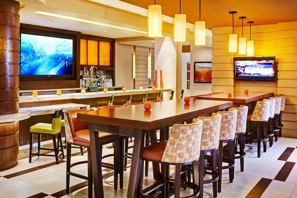 Upscale lounge with orange walls, bar seating, dining table, pendant lights, and multiple TV screens