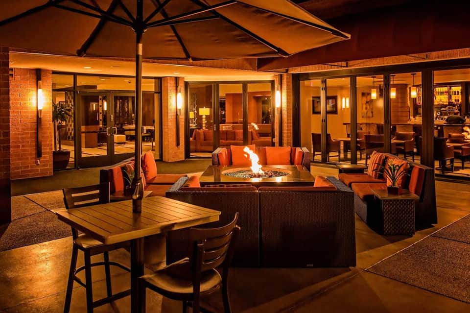 Evening patio with fire pit seating area, orange accent pillows, and modern furniture overlooking lit building