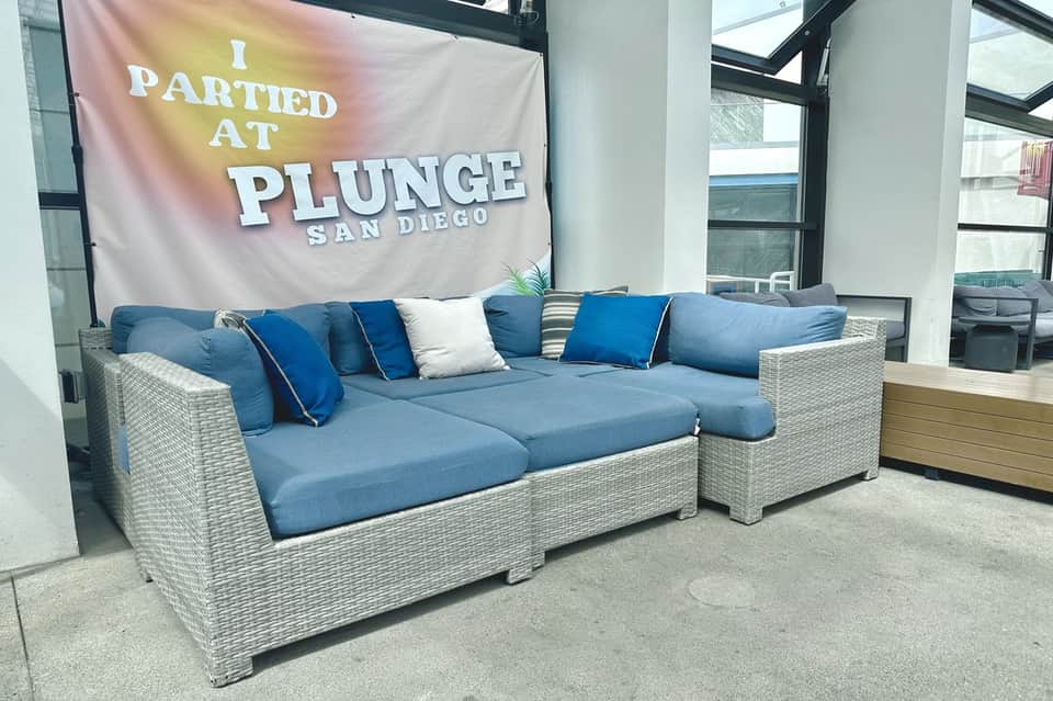 Modern rattan sectional sofa with blue cushions in hotel lobby under 'I Partied at Plunge San Diego' banner