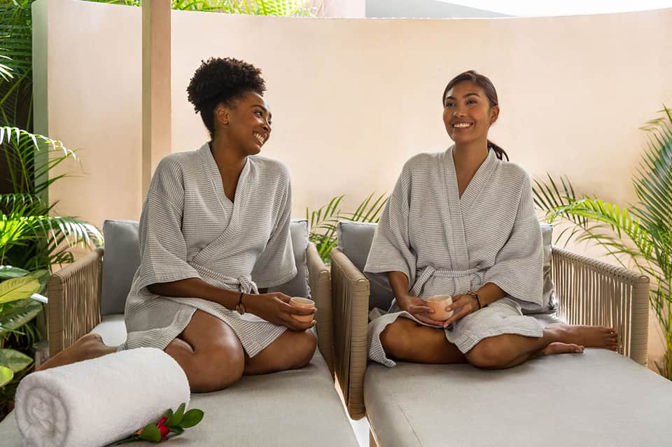 Two women in spa robes sitting on massage beds in a plant-filled wellness room