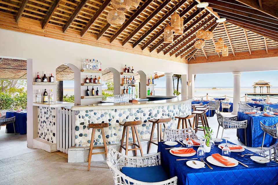 Beachfront restaurant with white bar featuring blue patterned facade, wooden ceiling, and ocean views with blue umbrellas