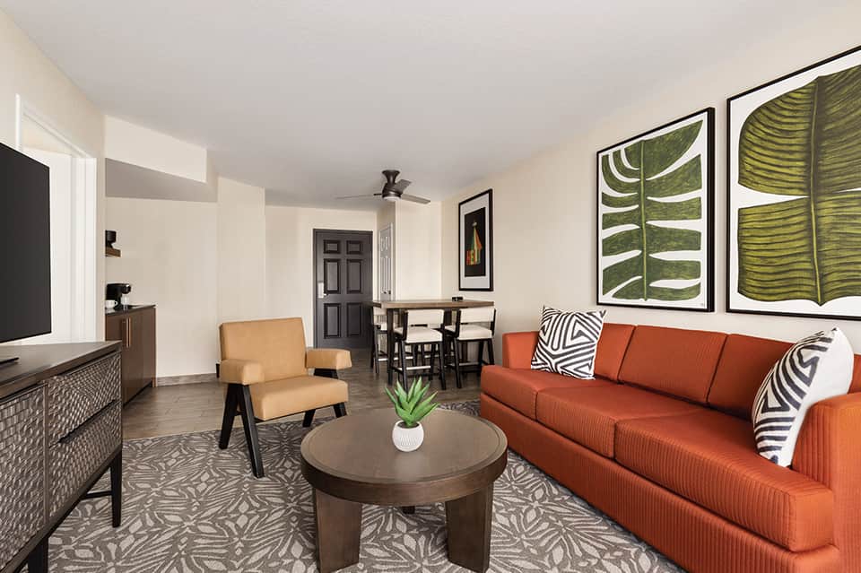 Modern hotel suite with orange sofa, tan armchair, dining area, and botanical wall art