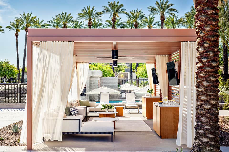 Covered outdoor lounge area with cream curtains, wooden pergola, lounge seating, and pool view surrounded by palm trees