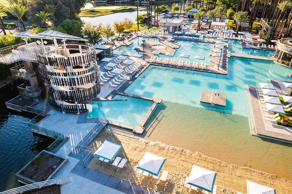 Aerial view of large resort pool complex with multiple pools, sunbathing areas, umbrellas, and tropical landscaping