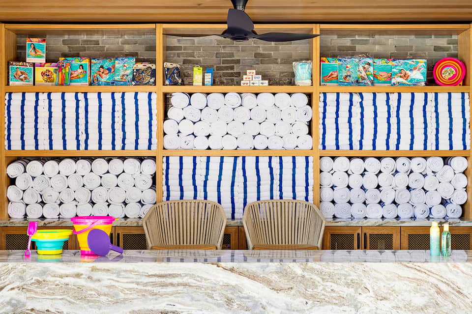 Beach cabana storage with rolled white towels, striped cushions, colorful beach toys, and surfboards on wooden shelves.
