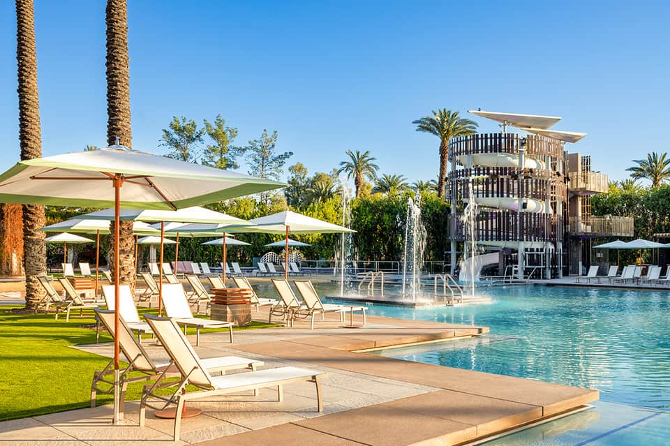 Luxury pool area with green umbrellas, lounge chairs, palm trees, and modern architectural water feature building