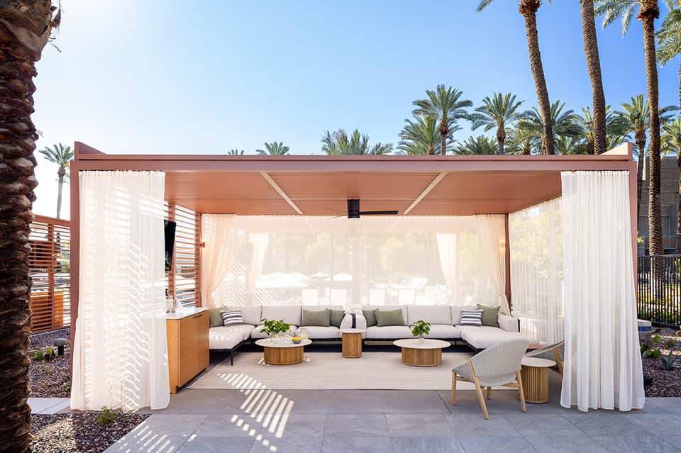 Luxury poolside cabana with sheer white curtains, wooden furniture, and palm trees in background
