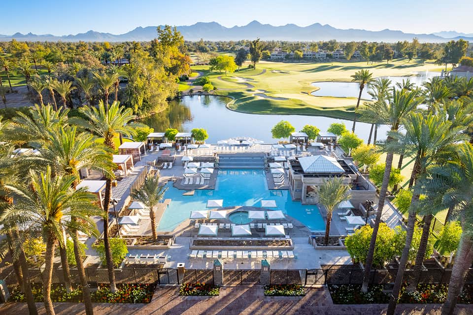 Aerial view of resort pool surrounded by palm trees with golf course and mountains in background