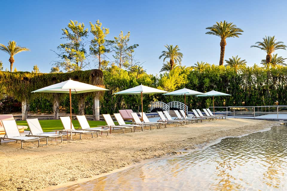 Beach resort with white umbrellas, lounge chairs, palm trees, and calm shallow water