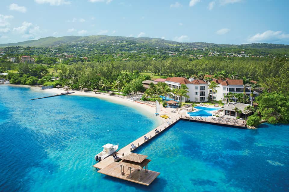 Aerial view of beachfront resort with white buildings, blue pool, and lush green hillside backdrop