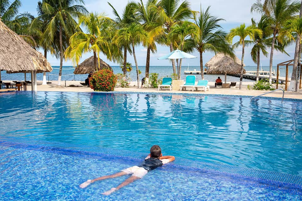 Resort swimming pool with tropical palm trees, thatched umbrellas, and ocean view in background