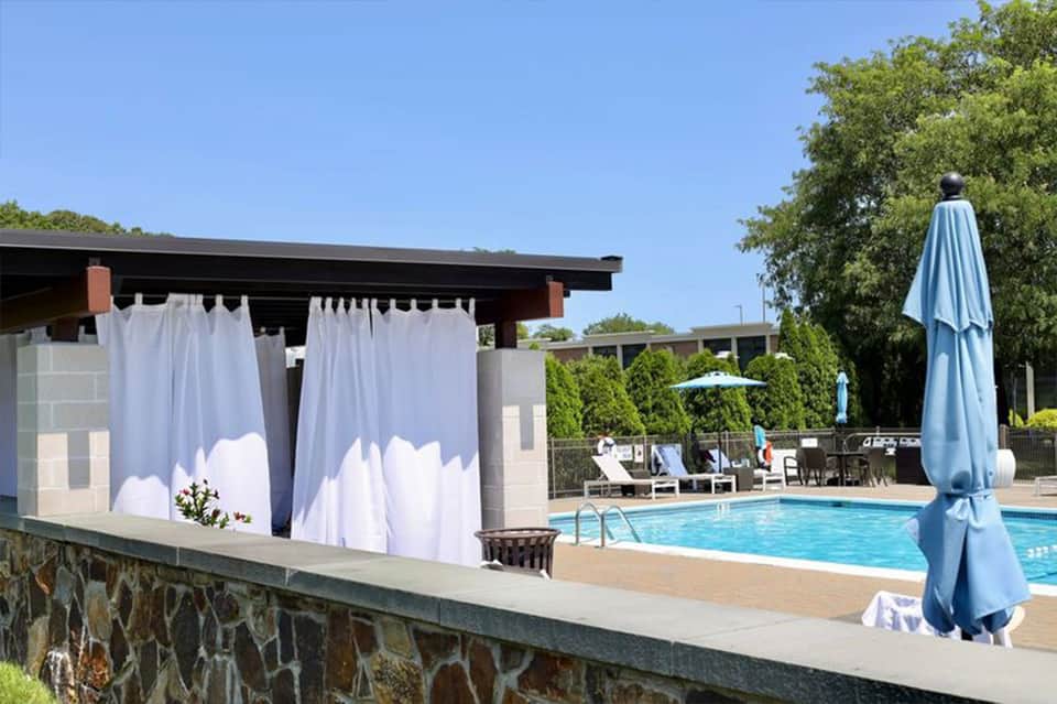 Resort pool area with white cabana curtains, loungers, and blue umbrellas under clear sky