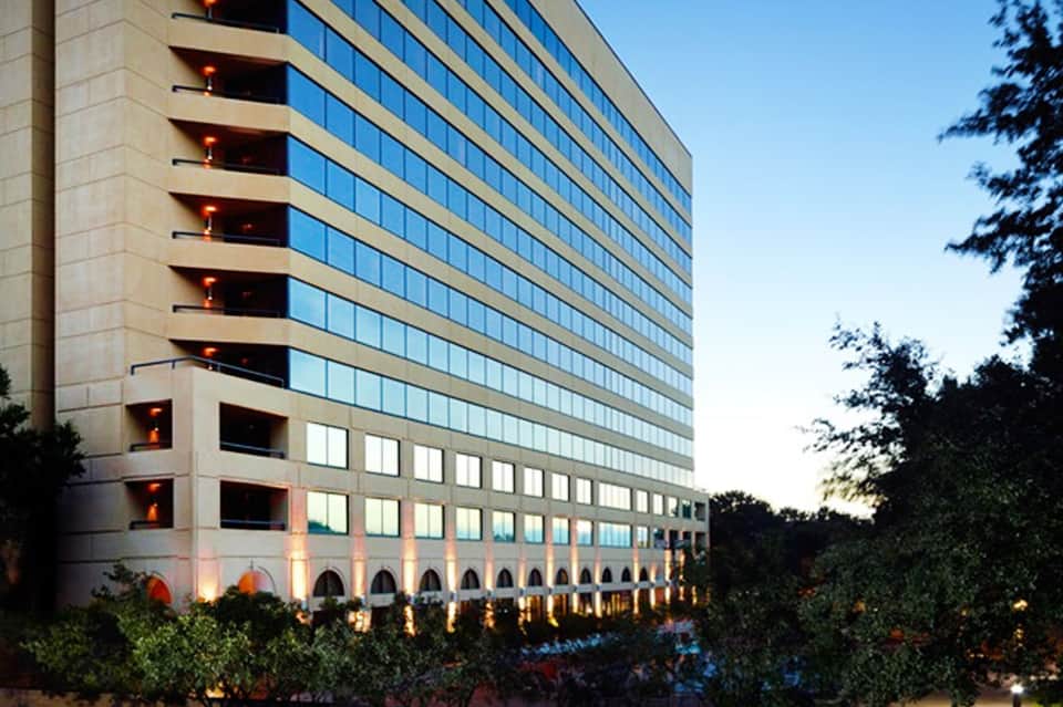 Modern multi-story hotel building with glass windows and lit balconies at dusk surrounded by landscaping