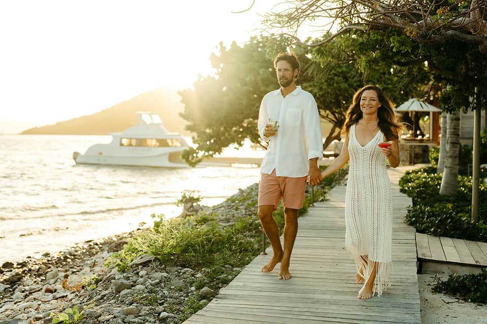 Couple walking on wooden boardwalk at sunset near beach with moored boat in background