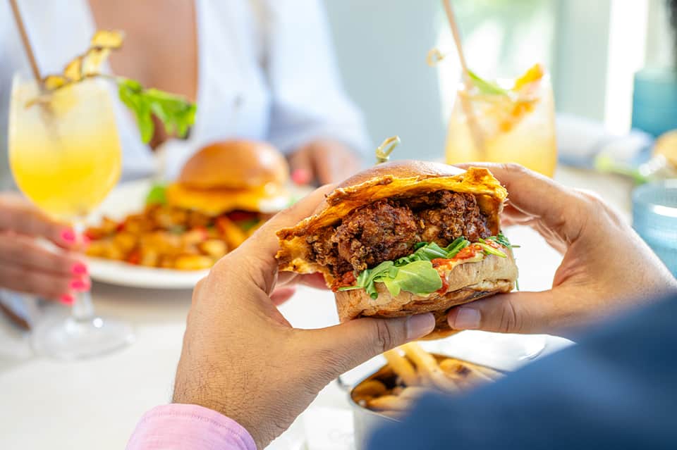 Hand holding a gourmet burger with fries and cocktails at an outdoor dining venue