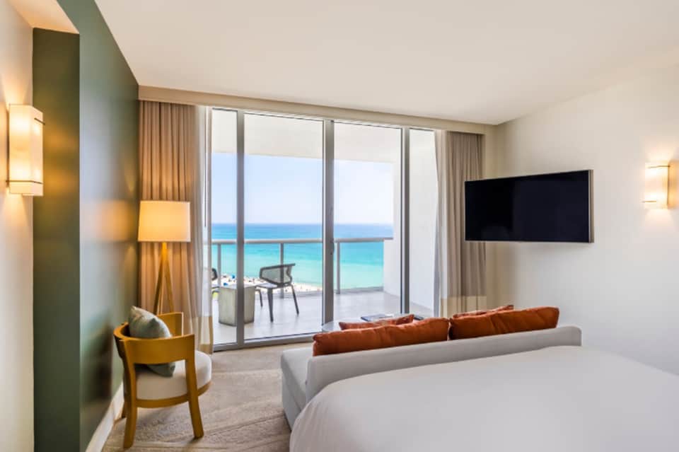 Luxury beachfront hotel room with ocean view, gray sofa, bed, and glass balcony doors opening to terrace