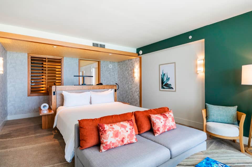 Modern hotel bedroom with green accent wall, wooden bed frame, and gray sofa with orange pillows