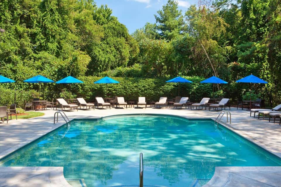 Resort pool with crystal blue water, lounge chairs, blue umbrellas, and lush green trees in background