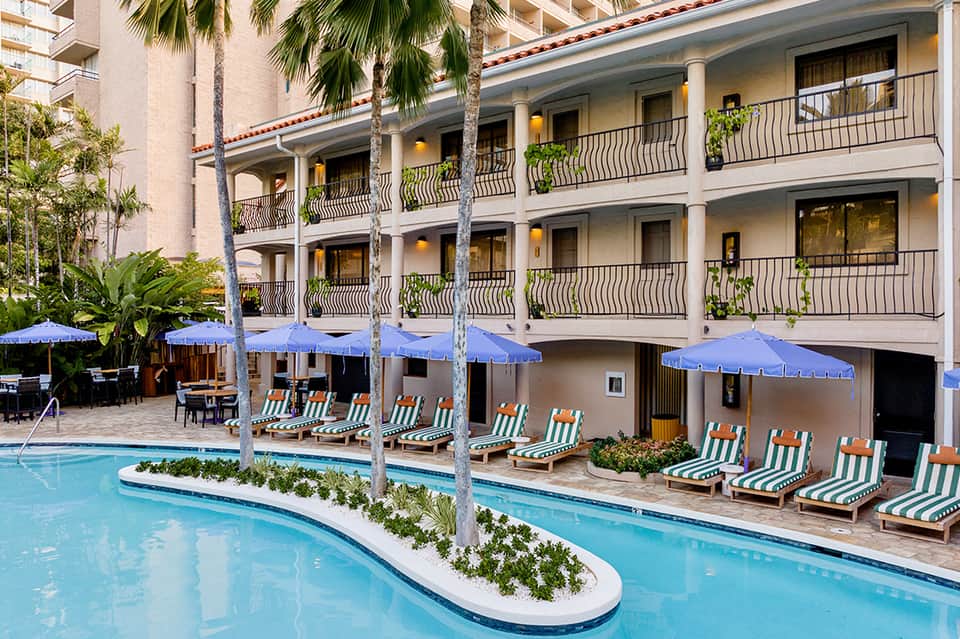 Multi-story hotel building with white columns, blue umbrellas, and striped lounge chairs by curved pool