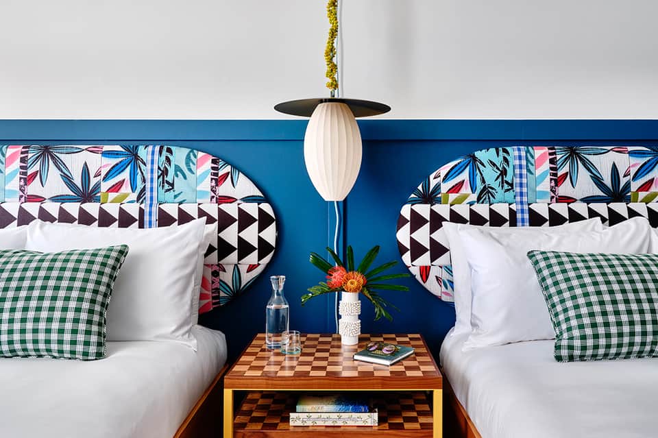 Modern hotel bedroom with twin beds, colorful patterned headboards, green gingham pillows, and pendant lighting