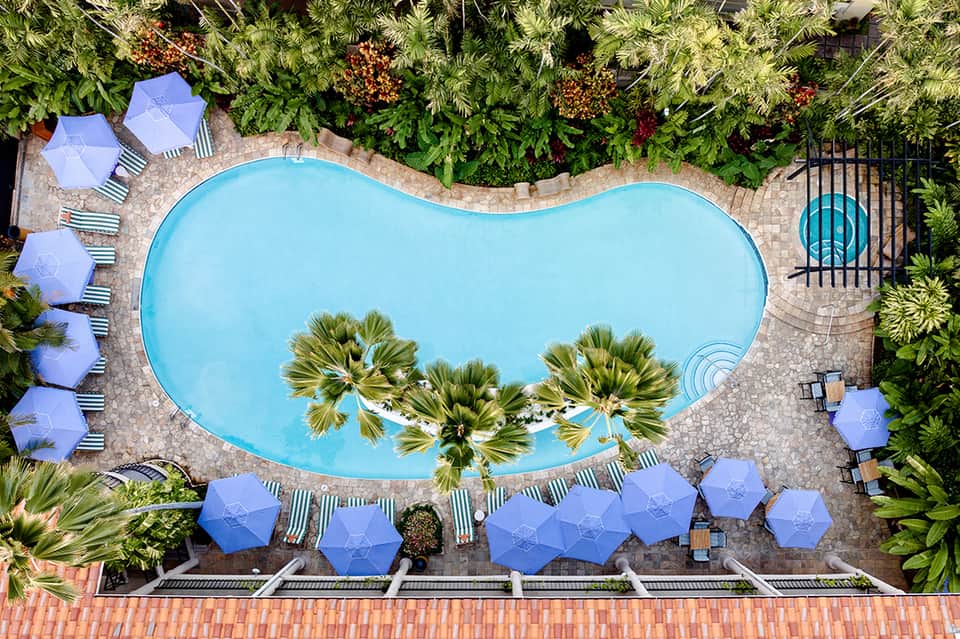 Aerial view of resort pool with blue umbrellas, lounge chairs, palm trees, and surrounding landscaping