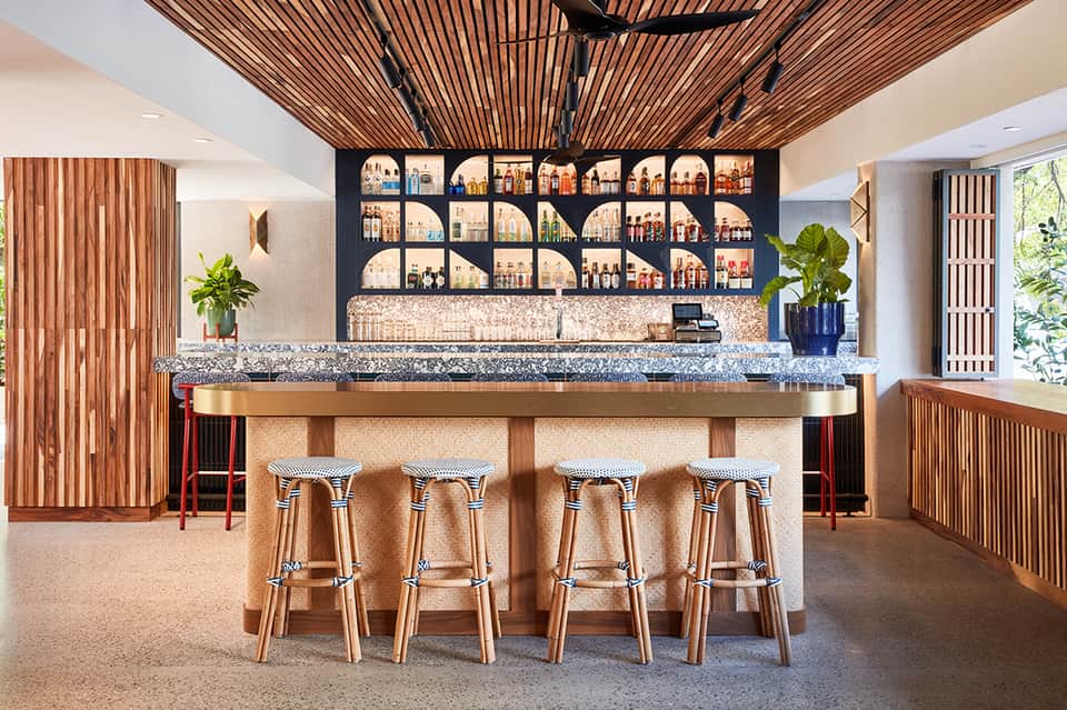 Modern bar interior with wooden slat ceiling, granite countertop, decorative shelving, and woven stools