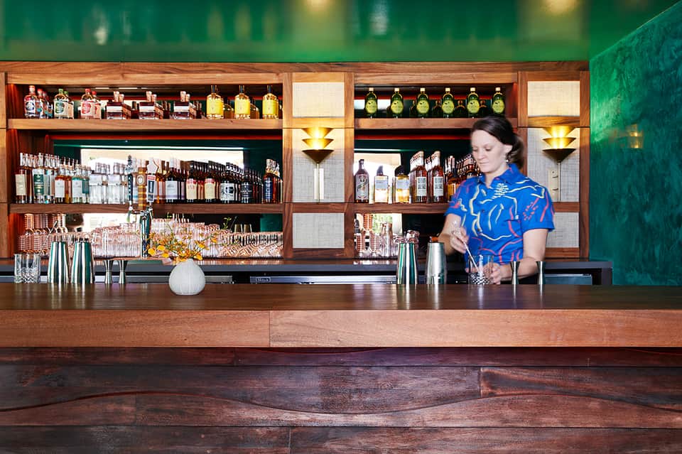 Bartender in blue patterned shirt preparing cocktails behind wooden bar with extensive liquor selection and green wall backdrop
