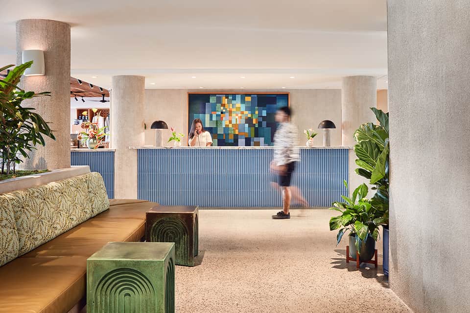 Modern hotel lobby with blue-paneled reception desk, geometric wall art, potted plants, and column supports