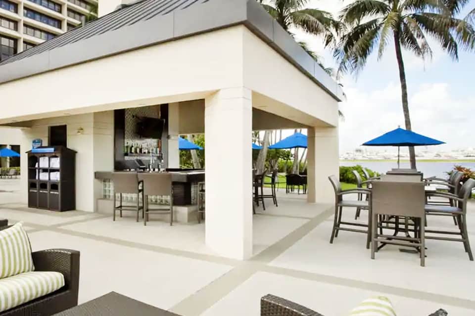 Modern beachfront patio with white lounge seating, bar area, umbrellas, and water views