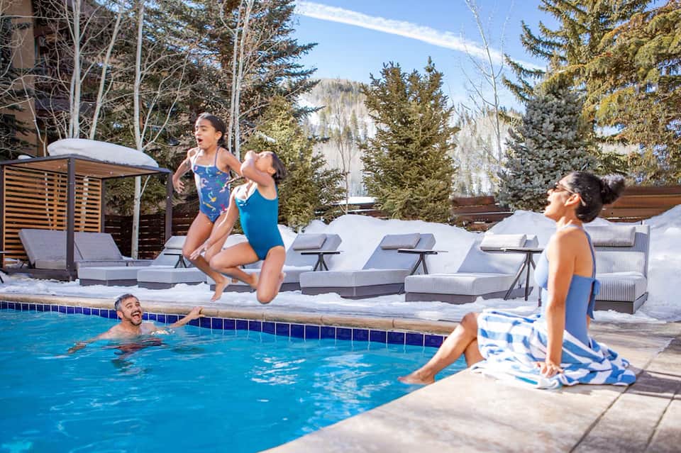 Family enjoying heated outdoor pool with snowy landscape and evergreen trees in winter mountain setting