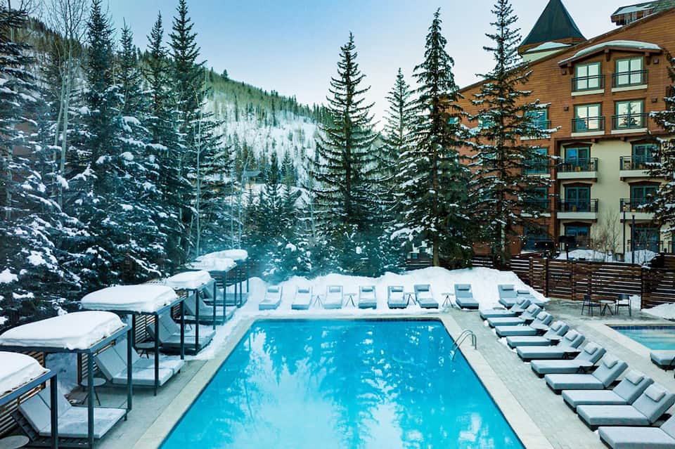 Mountain resort pool surrounded by snow-covered loungers with lodge building and pine forest backdrop